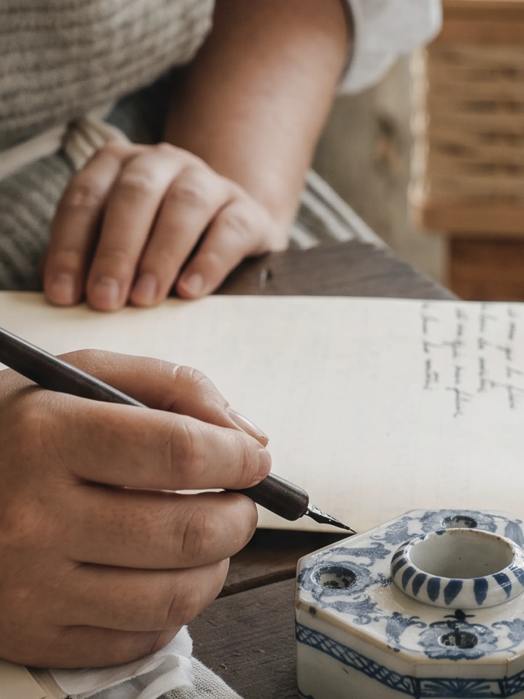 A hand writing with a fountain pen beside a blue-and-white ceramic inkwell.