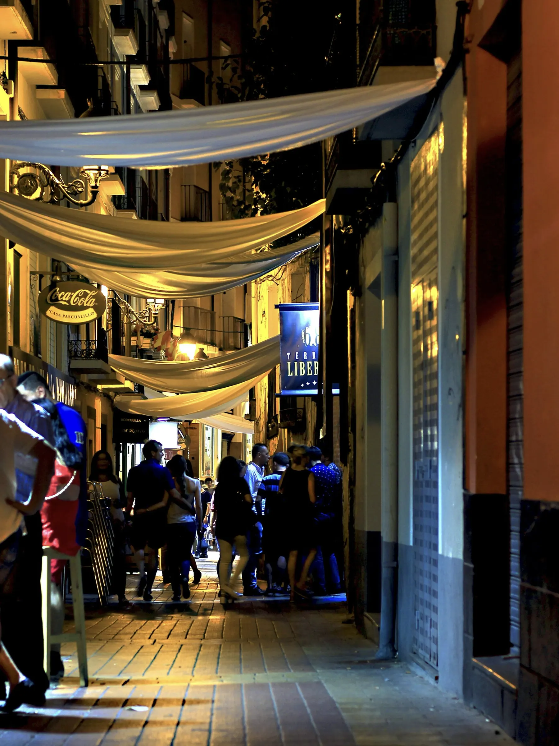 Evening in a narrow Spanish street strung with canvas awnings, people walking under the lights.