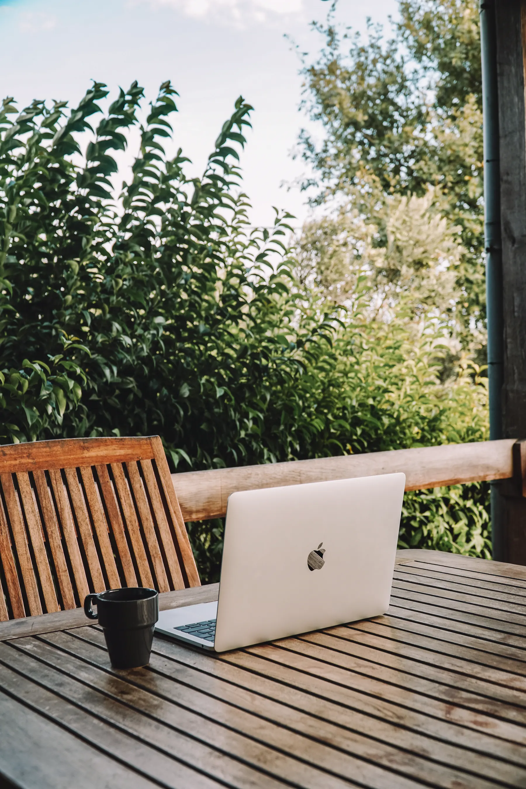 A laptop and a coffee cup on a wooden terrace table, green hedges behind.