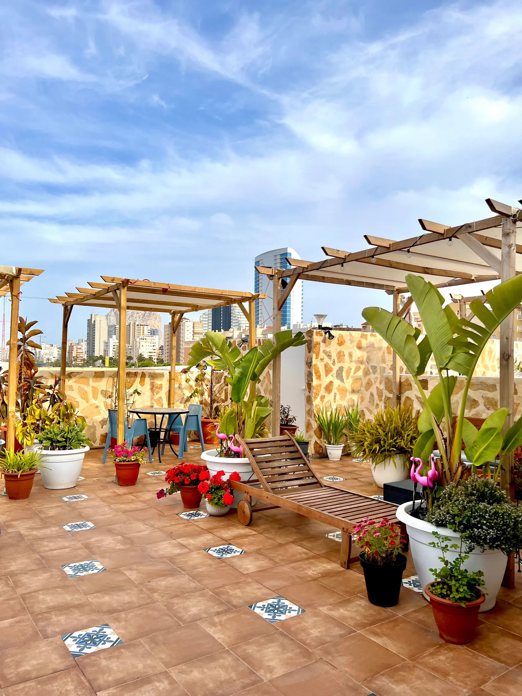 A sunlit roof terrace with timber pergolas, potted plants and patterned tiles, the city skyline beyond.