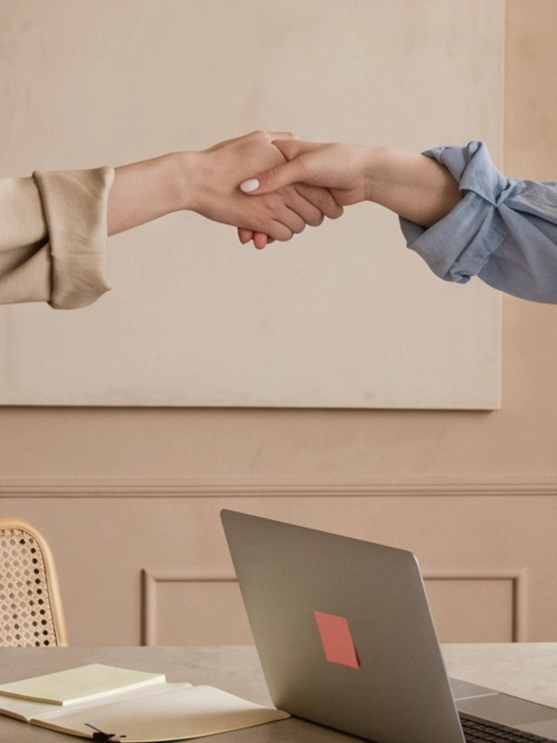 Two people shaking hands across a desk, a laptop and an open notebook in the foreground.