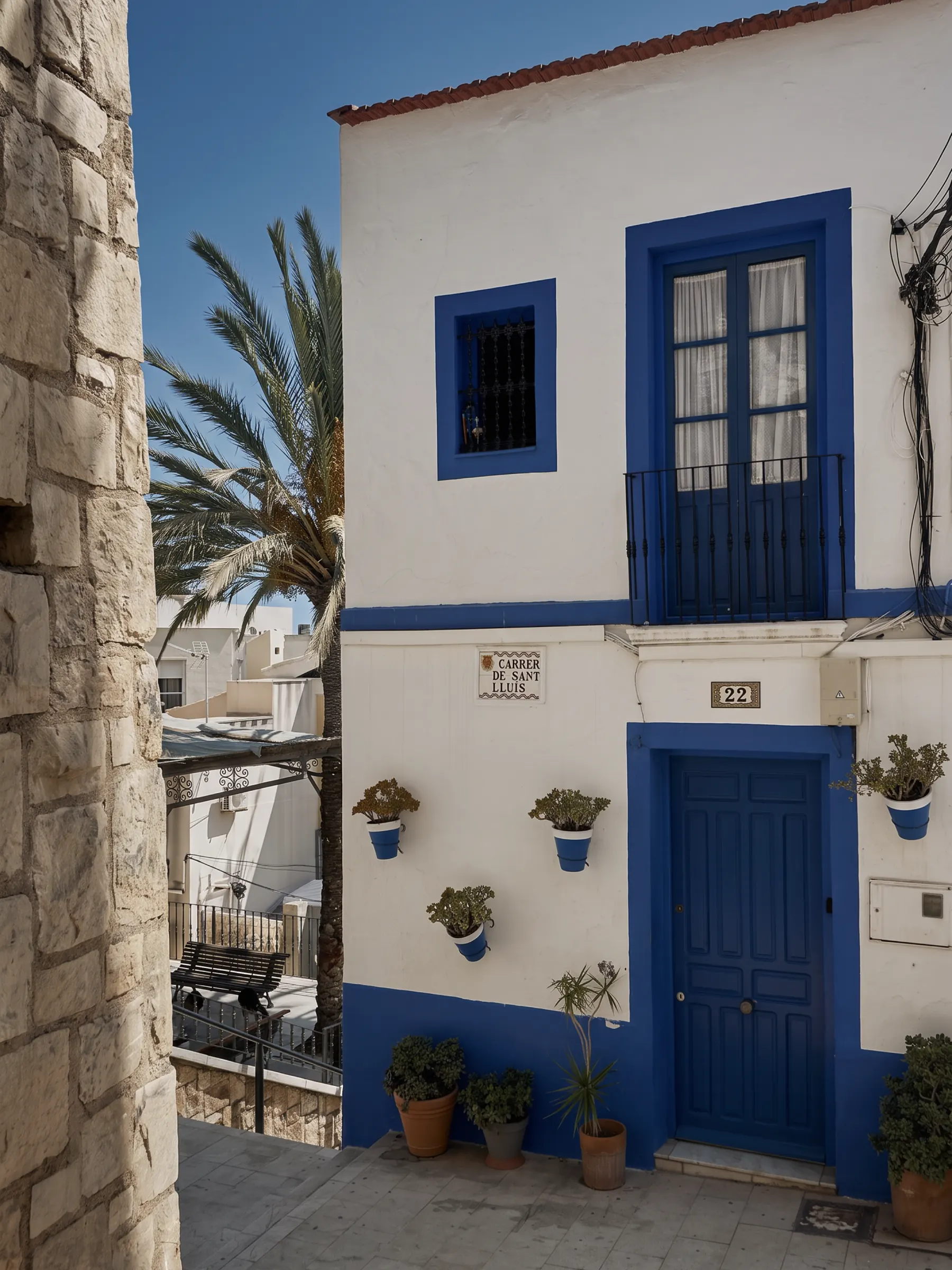 A whitewashed Mediterranean house with a cobalt blue door and shutters, potted geraniums on the wall.